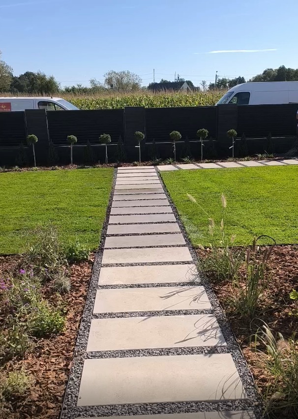 Nowoczesny ogród z trawnikiem, ścieżką z płyt i czarnym ogrodzeniem. Małe drzewka kuliste wzdłuż ogrodzenia. W tle budynki i zieleń.