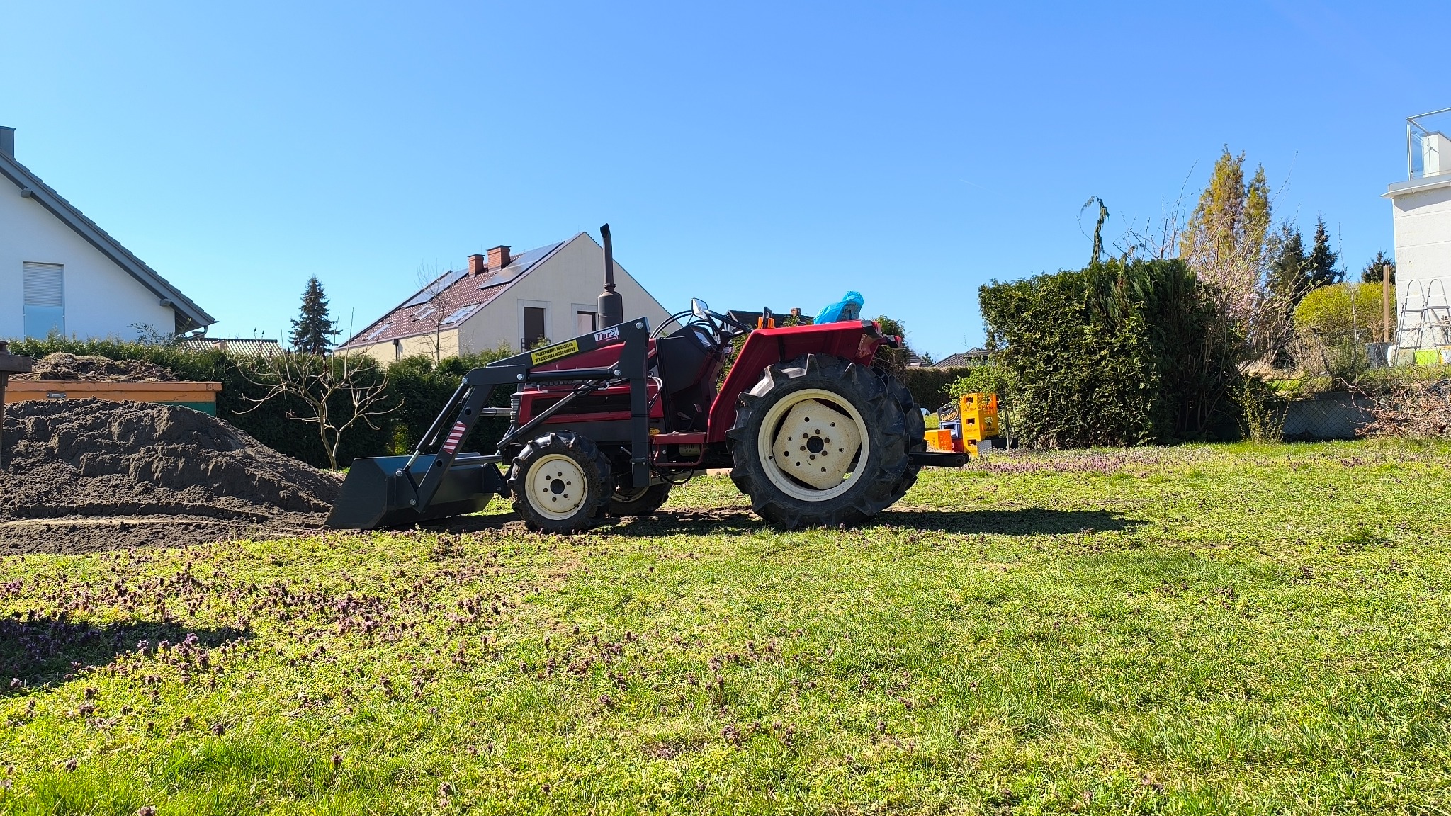 Czerwony traktor z łyżką na trawniku przed domem, obok pryzma ziemi. Widoczne budynki w tle i błękitne niebo. Prace ziemne w ogrodzie.