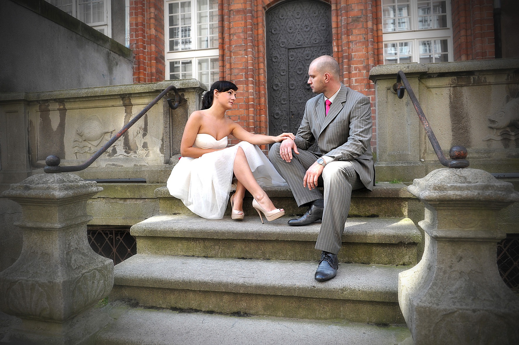 Para młoda siedząca na kamiennych schodach przed budynkiem z czerwonej cegły; panna młoda w białej sukni bez ramiączek i szpilkach, pan młody w szarym garniturze w prążki i różowym krawacie.