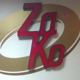 Logo firmy ZaKo: czerwone litery na złotym owalu, umieszczone na białej ścianie. Widoczny znak zastrzeżony 'R' w kółku.