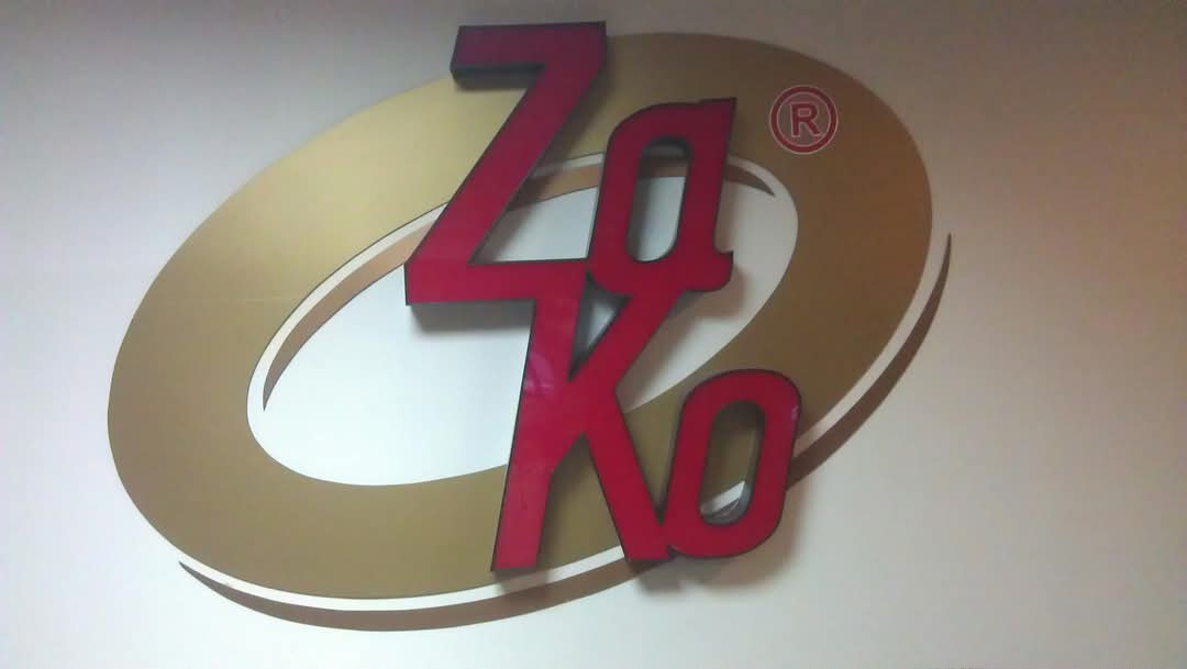 Logo firmy ZaKo: czerwone litery na złotym owalu, umieszczone na białej ścianie. Widoczny znak zastrzeżony 'R' w kółku.