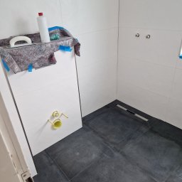 Majer-bau - Nowa łazienka z ciemnymi płytkami podłogowymi i jasnymi ścianami, widoczny odpływ liniowy, przygotowane przyłącza do baterii, narzędzia i preparaty zabezpieczone folią malarską.