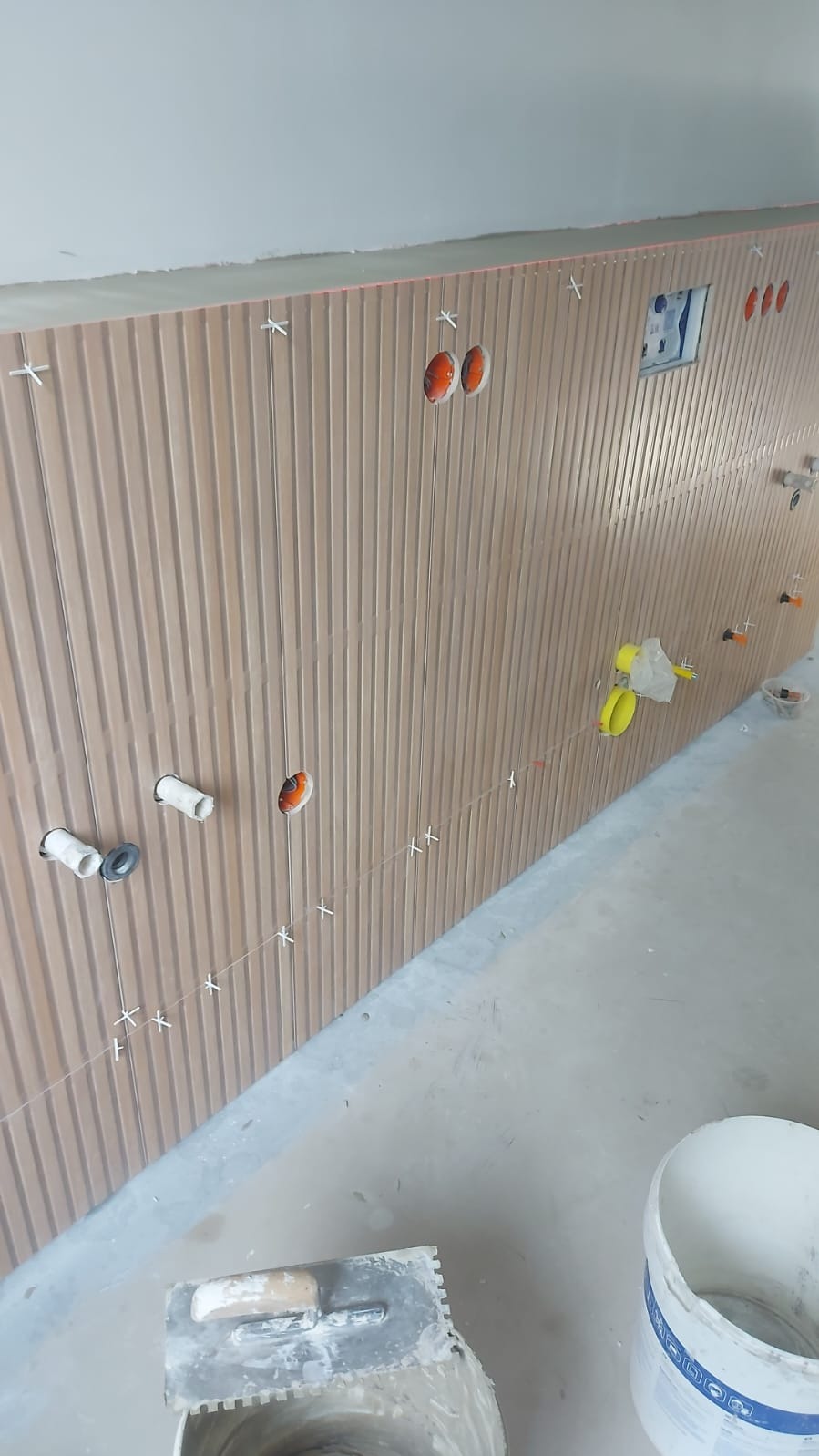 Ściana wyłożona pionowymi, brązowymi płytkami imitującymi lamele, z widocznymi otworami na instalacje i krzyżykami dystansowymi, w trakcie prac glazurniczych.