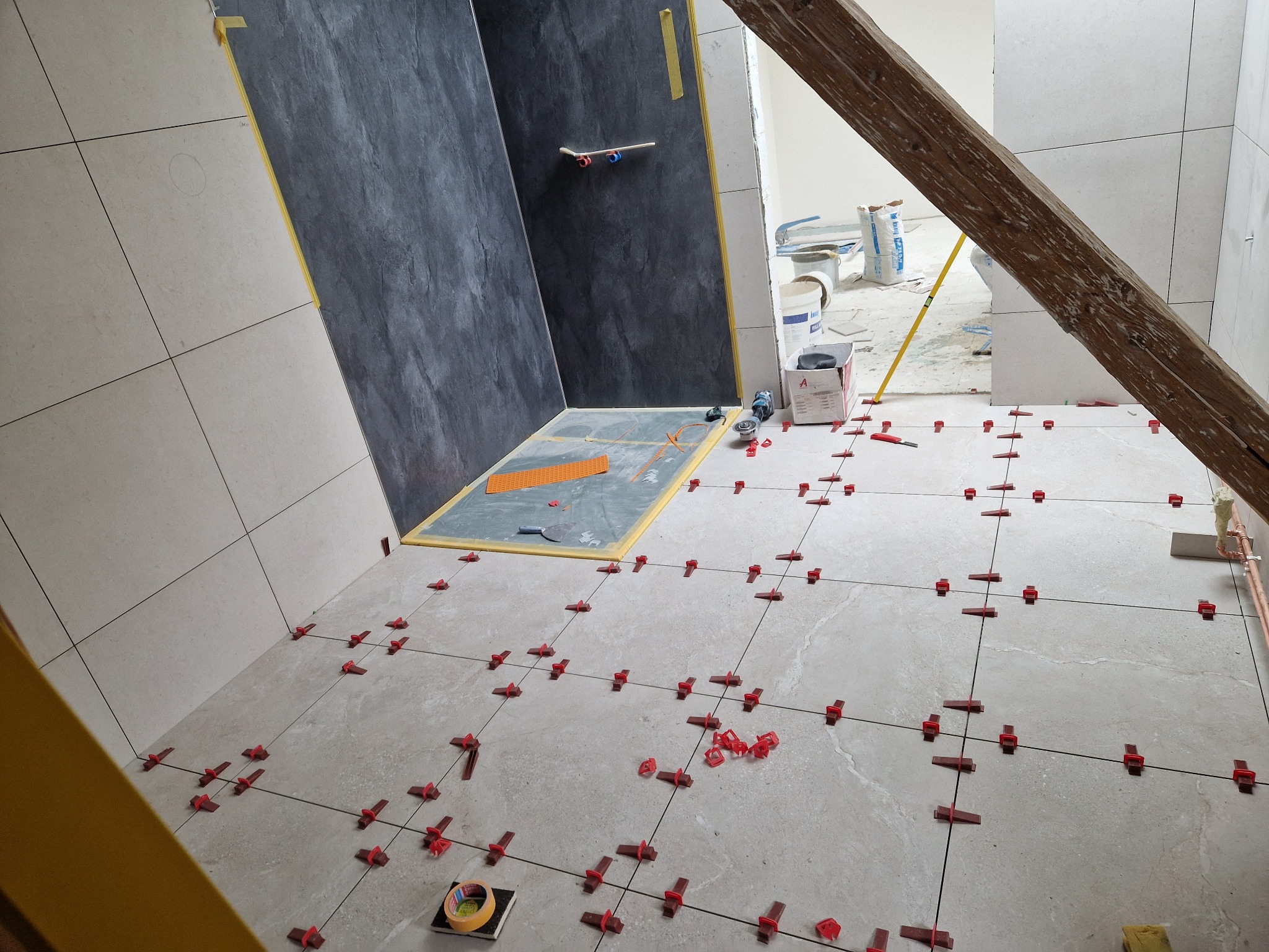 Układanie płytek podłogowych w Miastko z użyciem poziomowania, widoczne krzyżyki dystansowe i fragment ściany z ciemnymi płytkami imitującymi kamień.