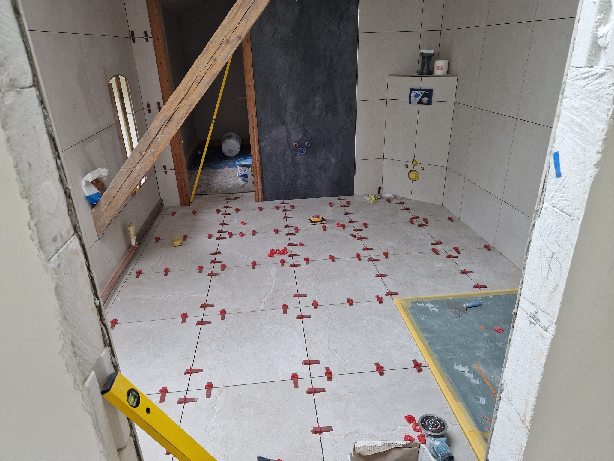 Łazienka w trakcie układania jasnych płytek podłogowych, z widocznymi krzyżykami dystansowymi, poziomica przy ościeżnicy, oraz fragment ściany w ciemnej kolorystyce.