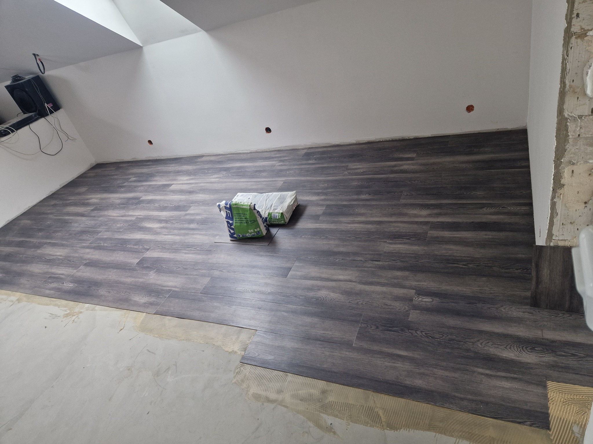 Układanie paneli podłogowych w poddaszu, widoczne worki z klejem i fragment ściany w stanie surowym.