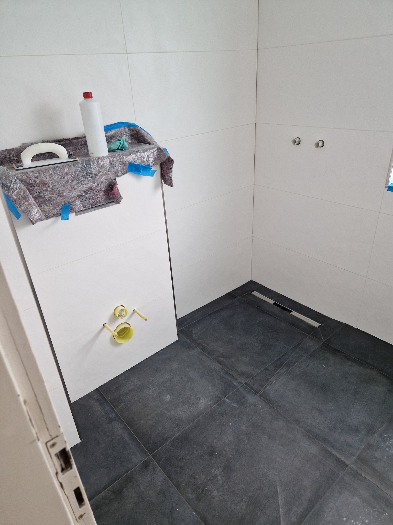 Nowa łazienka z ciemnymi płytkami podłogowymi i jasnymi ścianami, widoczny odpływ liniowy, przygotowane przyłącza do baterii, narzędzia i preparaty zabezpieczone folią malarską.
