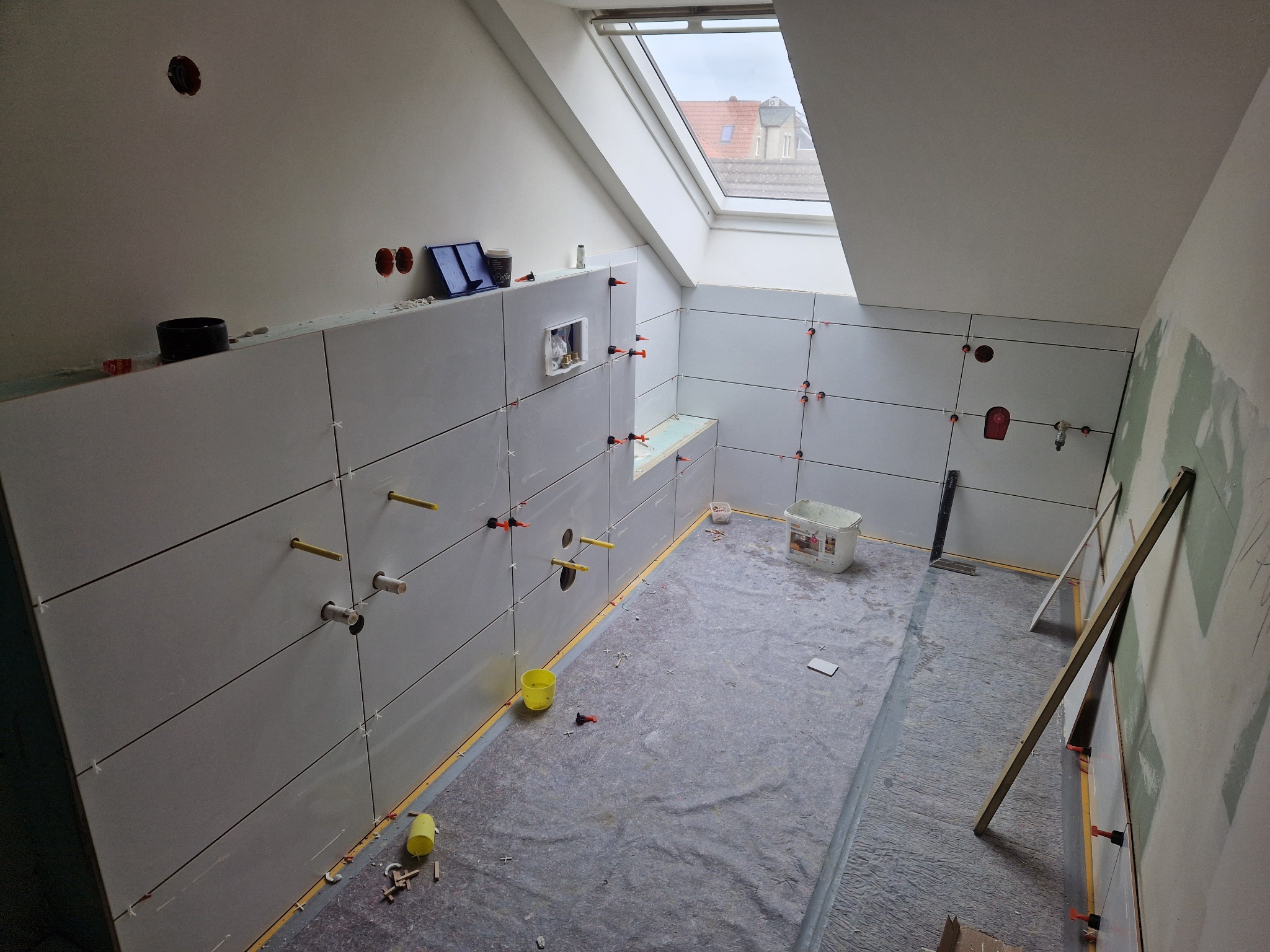Białe płytki ścienne w trakcie układania w łazience na poddaszu, widoczne poziomice, plastikowe kliny i otwory na instalacje, podłoga zabezpieczona folią ochronną.