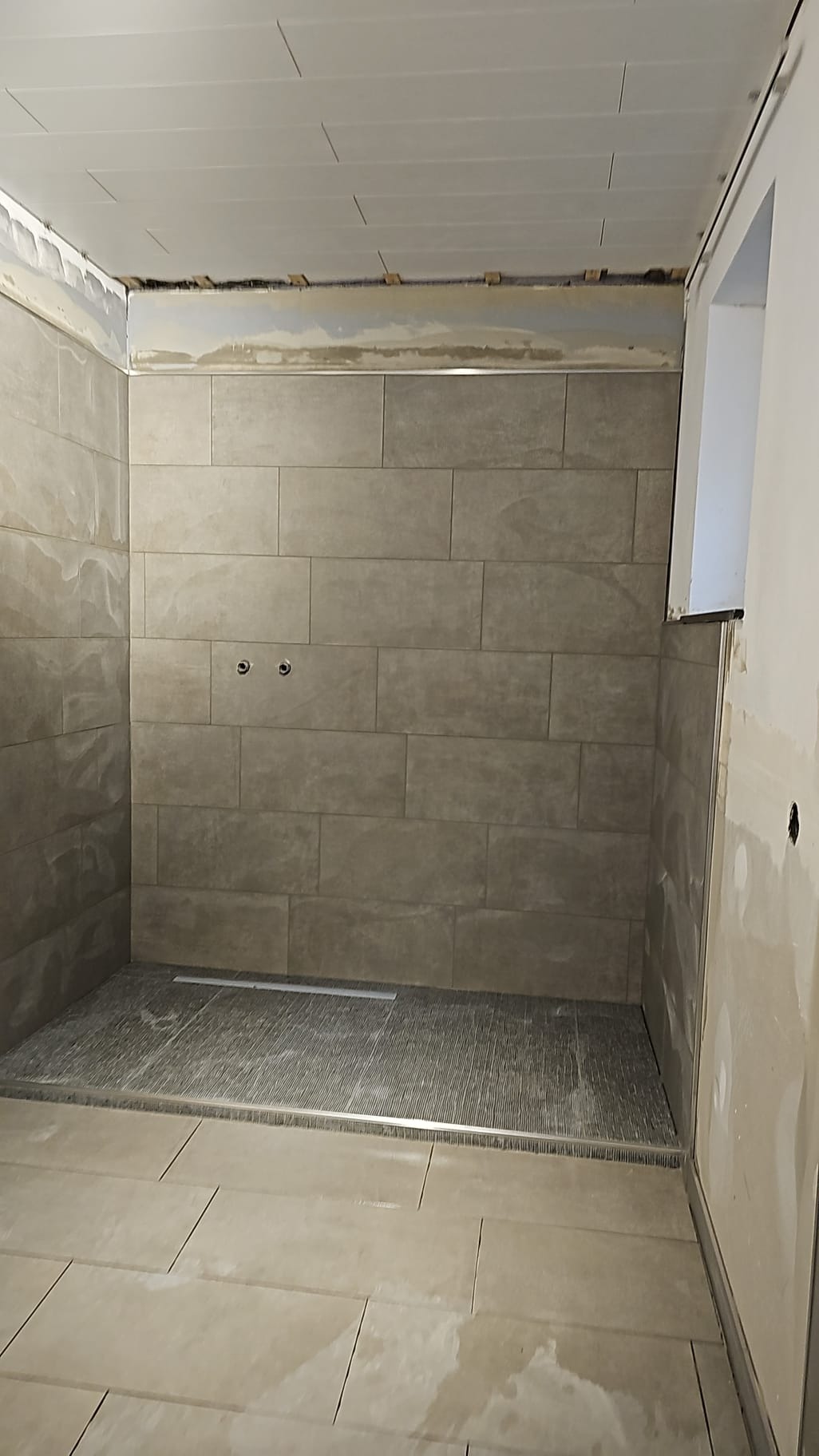 Wnętrze łazienki w trakcie prac wykończeniowych: ułożone beżowe płytki ścienne i podłogowe, widoczny odpływ liniowy, surowy sufit z widocznymi elementami konstrukcyjnymi.