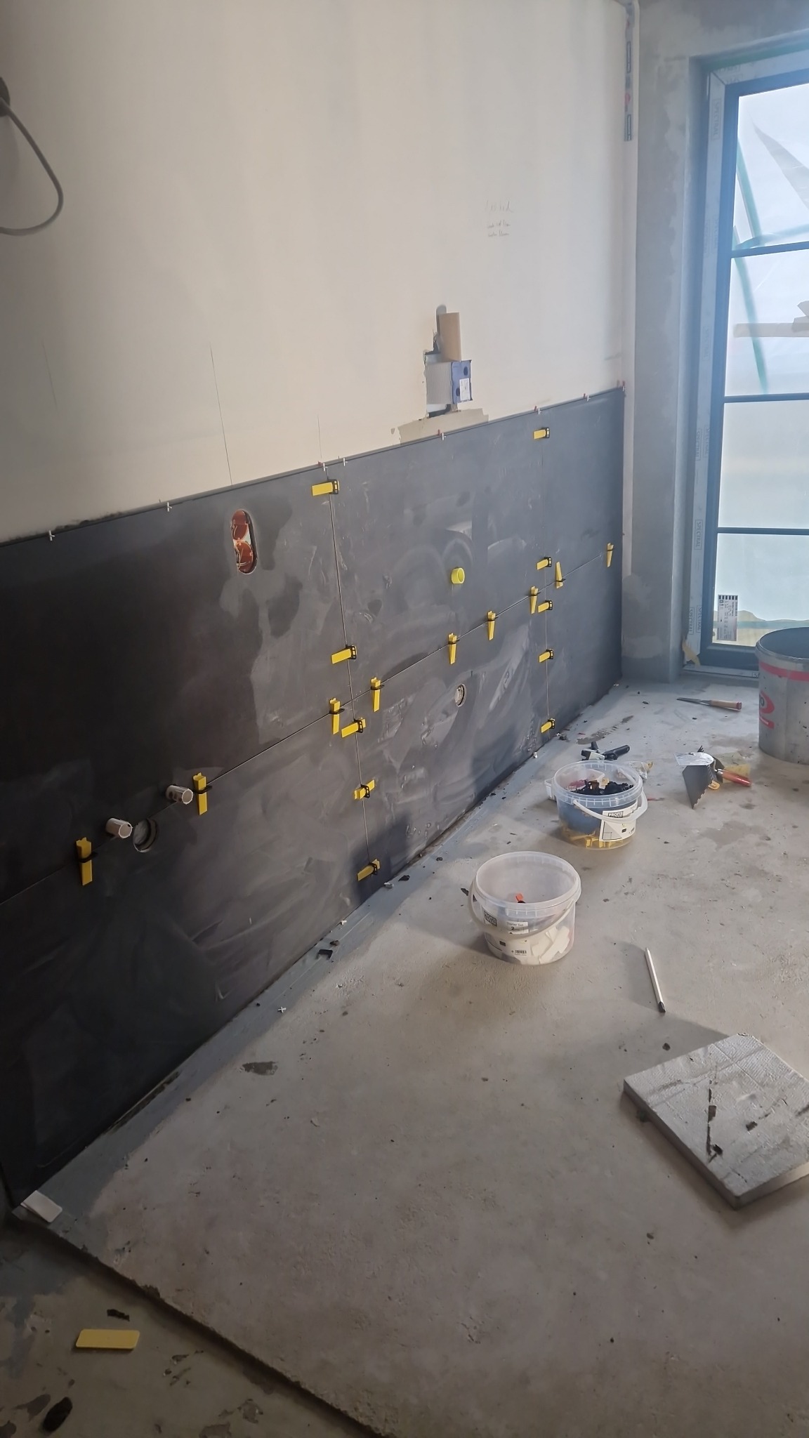 Ściana pokryta ciemnymi płytkami w trakcie montażu, widoczne żółte krzyżyki dystansowe, puszki elektryczne, rury, wiaderka z zaprawą i narzędzia na szarym, surowym podłożu.