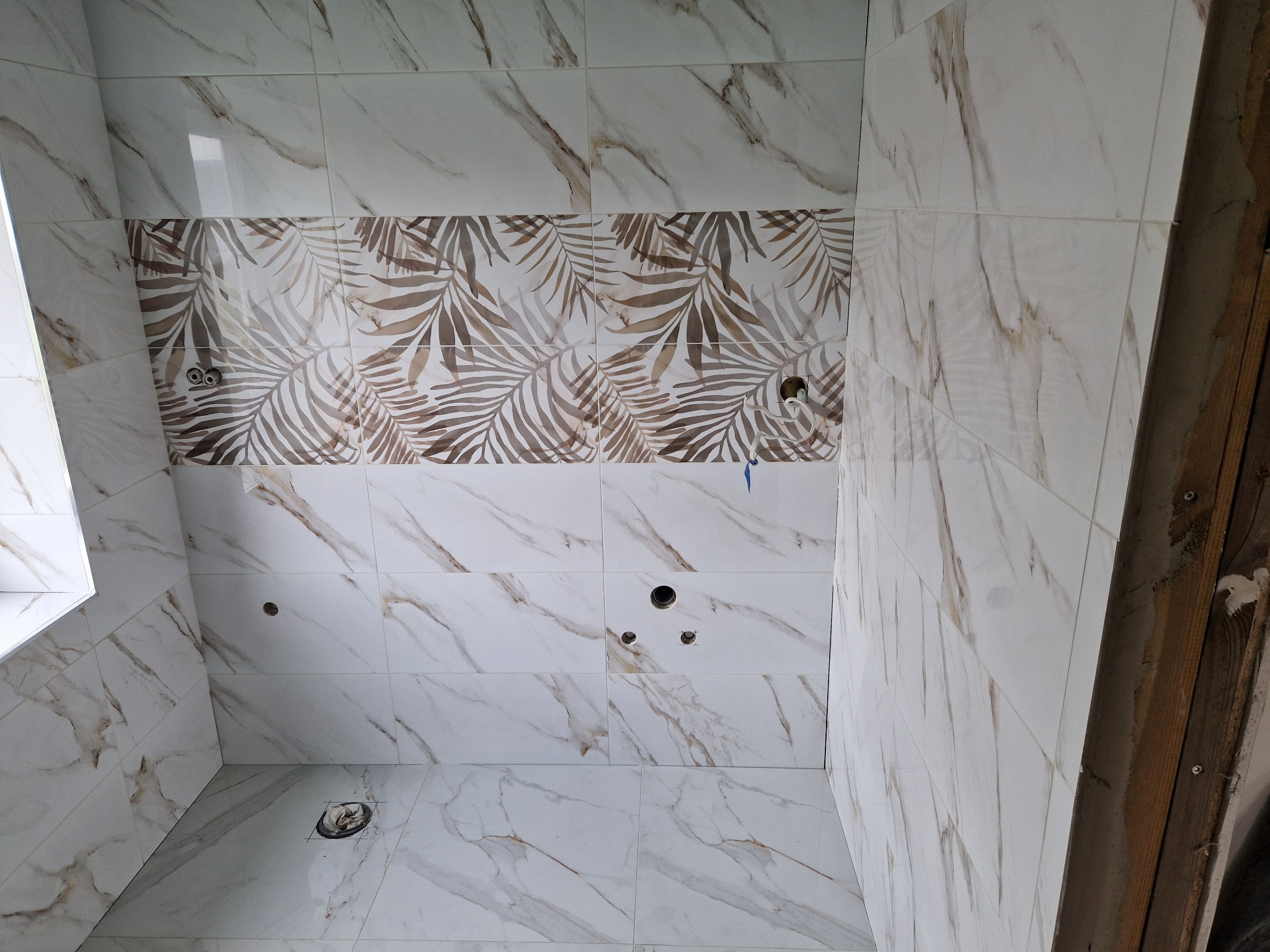 Ściany i podłoga łazienki wyłożone białymi płytkami imitującymi marmur z brązowymi żyłami, z poziomym pasem dekoracyjnych płytek z motywem brązowych liści palmowych, widoczne otwory na armaturę.