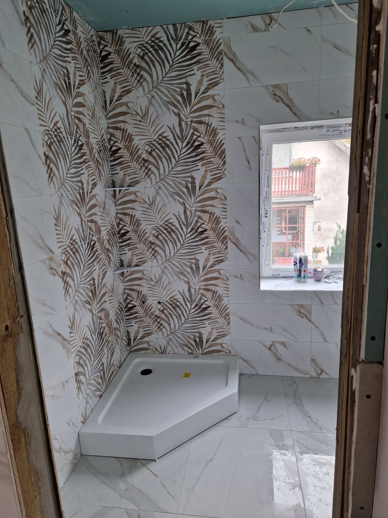 Niedokończona łazienka z zamontowanym brodzikiem, marmurowe płytki z motywem liści palmowych na ścianie, widoczne narzędzia i materiały budowlane na parapecie okiennym.