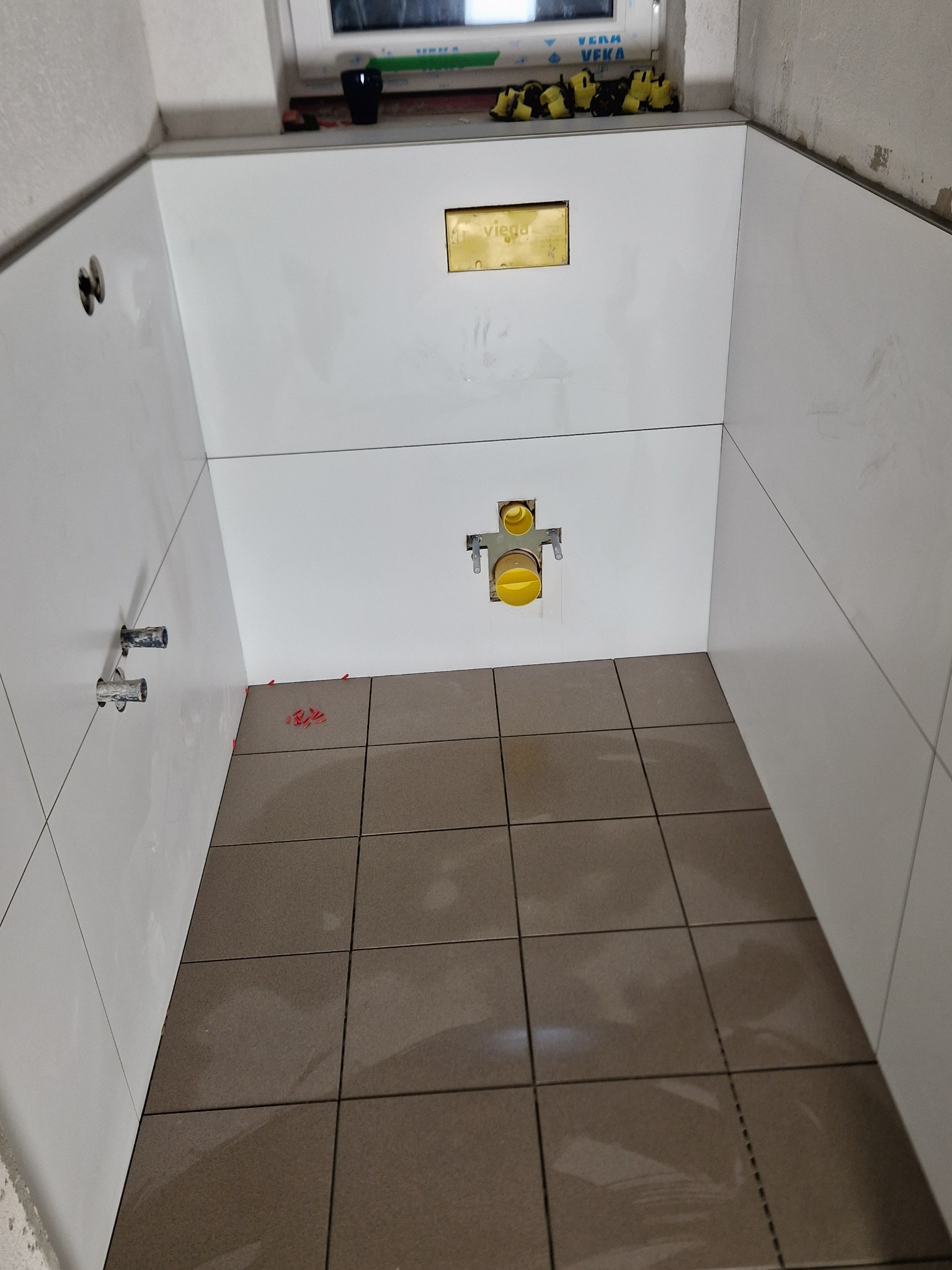 Białe płytki ścienne i brązowe płytki podłogowe w trakcie układania, widoczne elementy instalacji sanitarnej i poziomica.