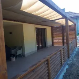 Zadaszony taras z drewnianą konstrukcją i materiałowym dachem, widoczne drewniane balustrady i ażurowe panele boczne, w tle dom z ogrodem.