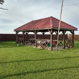 Drewniana altana z czerwonym dachem na zielonym trawniku, otoczona brązowym ogrodzeniem i huśtawką w tle.