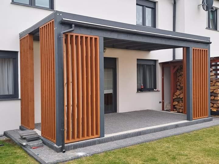 Nowoczesna pergola z drewnianymi lamelami w kolorze miodowym, z szarą konstrukcją, przylegająca do jasnego budynku z widocznymi oknami i stosem drewna w niszy, na tle zielonego trawnika.