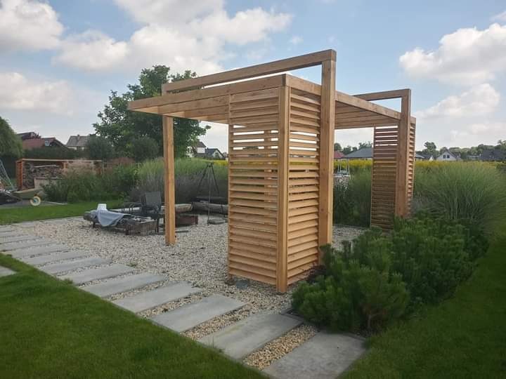 Drewniana konstrukcja z lamelami w ogrodzie, widok na palenisko i trawy ozdobne, ścieżka z płyt betonowych.