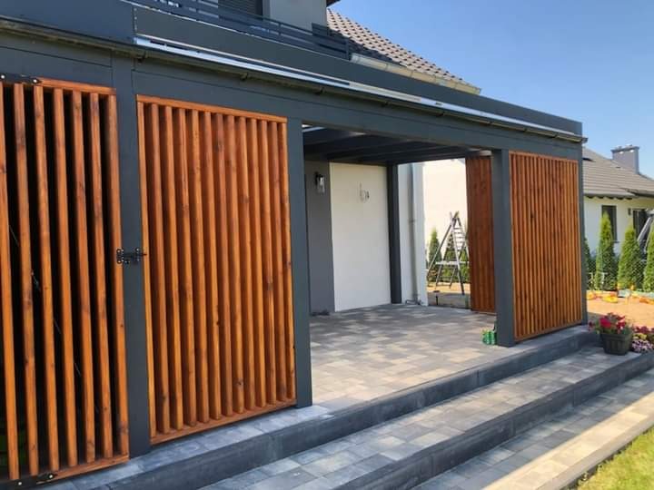 Nowoczesna pergola z drewnianymi panelami pionowymi w kolorze ciepłego brązu, osłonięta szarą konstrukcją, prowadząca do utwardzonego patio z widokiem na zadbany ogród.