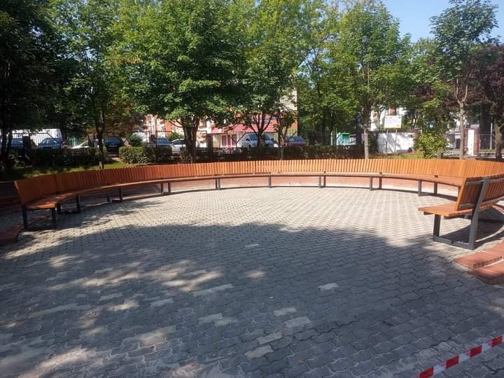 Półokrągła ławka z jasnego drewna i metalową konstrukcją, otaczająca plac wyłożony szarą kostką brukową, w tle drzewa i budynki.