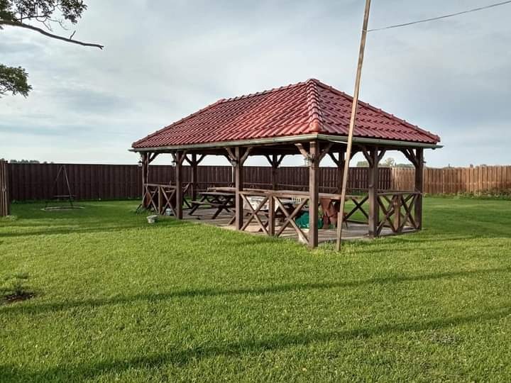 Drewniana altana z czerwonym dachem na zielonym trawniku, otoczona brązowym ogrodzeniem i huśtawką w tle.