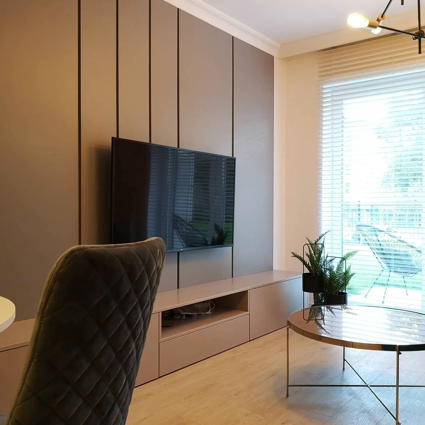 Nowoczesny salon z zabudową ściany w odcieniach beżu i brązu, telewizor, niska szafka RTV, okrągły stolik kawowy ze złotymi nogami i rośliny w doniczkach przy oknie.