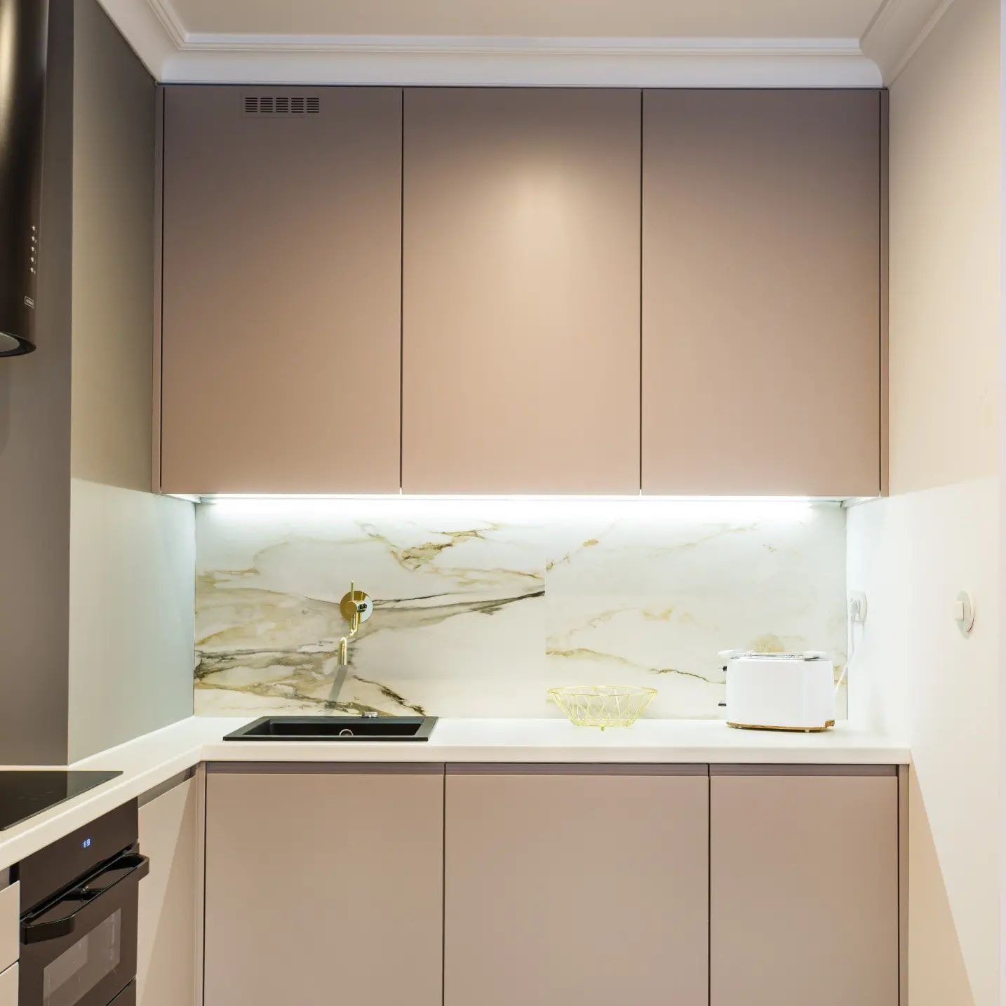 Nowoczesna kuchnia z gładkimi, beżowymi frontami szafek, białym blatem i marmurowym panelem ściennym podświetlonym listwą LED, czarny zlew i bateria w kolorze złotym.