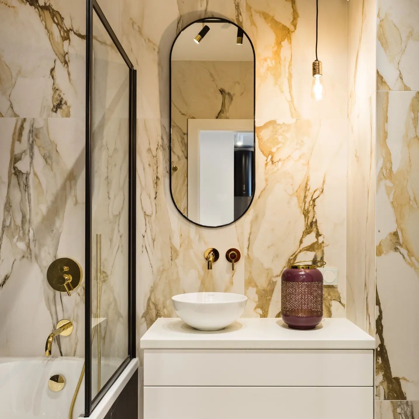 Elegancka łazienka z marmurowymi płytkami, złotą armaturą, owalnym lustrem z oświetleniem i białą szafką umywalkową z dekoracyjną fioletową latarenką.