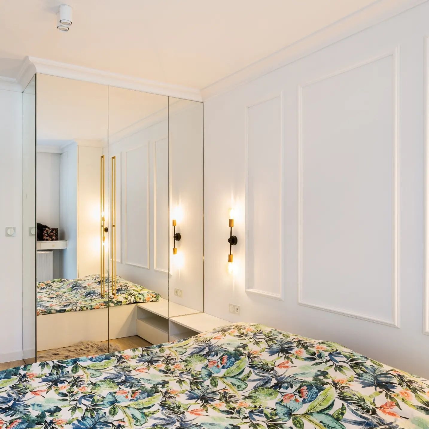 Biała szafa wnękowa z lustrzanymi drzwiami, wbudowana w ścianę z dekoracyjnymi panelami, oświetlona złotymi lampami, odbijająca wzór kwiatowy na pościeli.