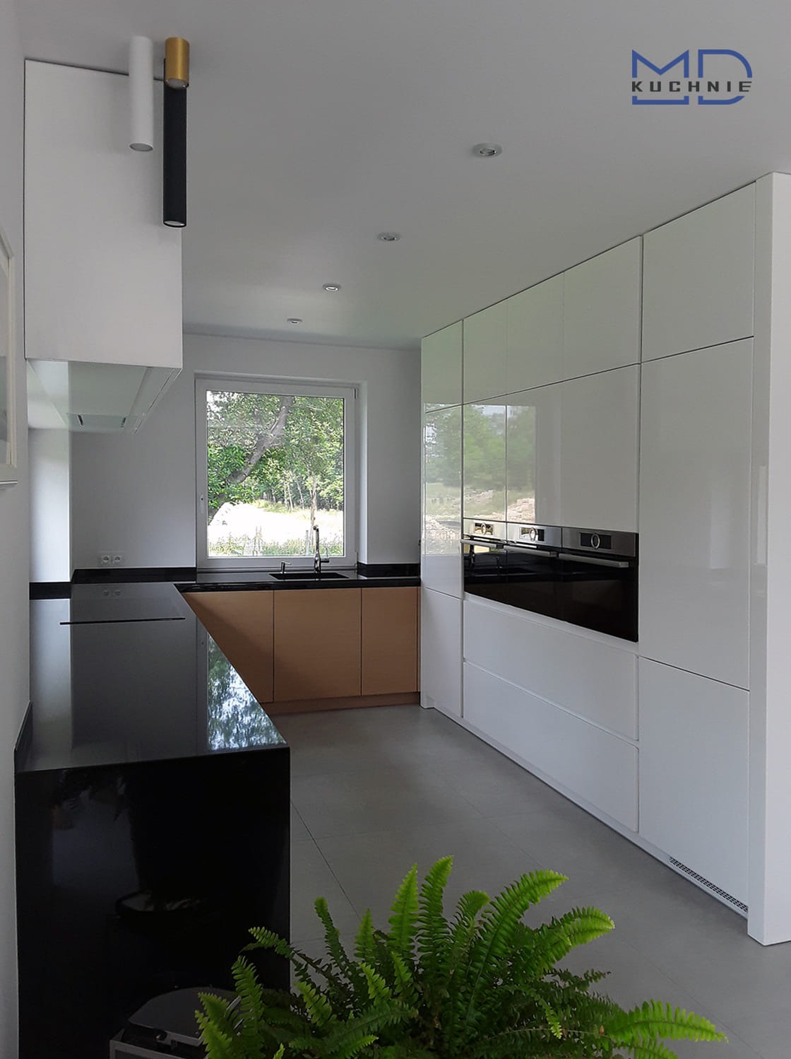 Nowoczesna kuchnia z białymi, lakierowanymi szafkami, czarnym blatem i drewnianymi frontami pod oknem, uzupełniona o zieloną roślinę doniczkową w rogu.