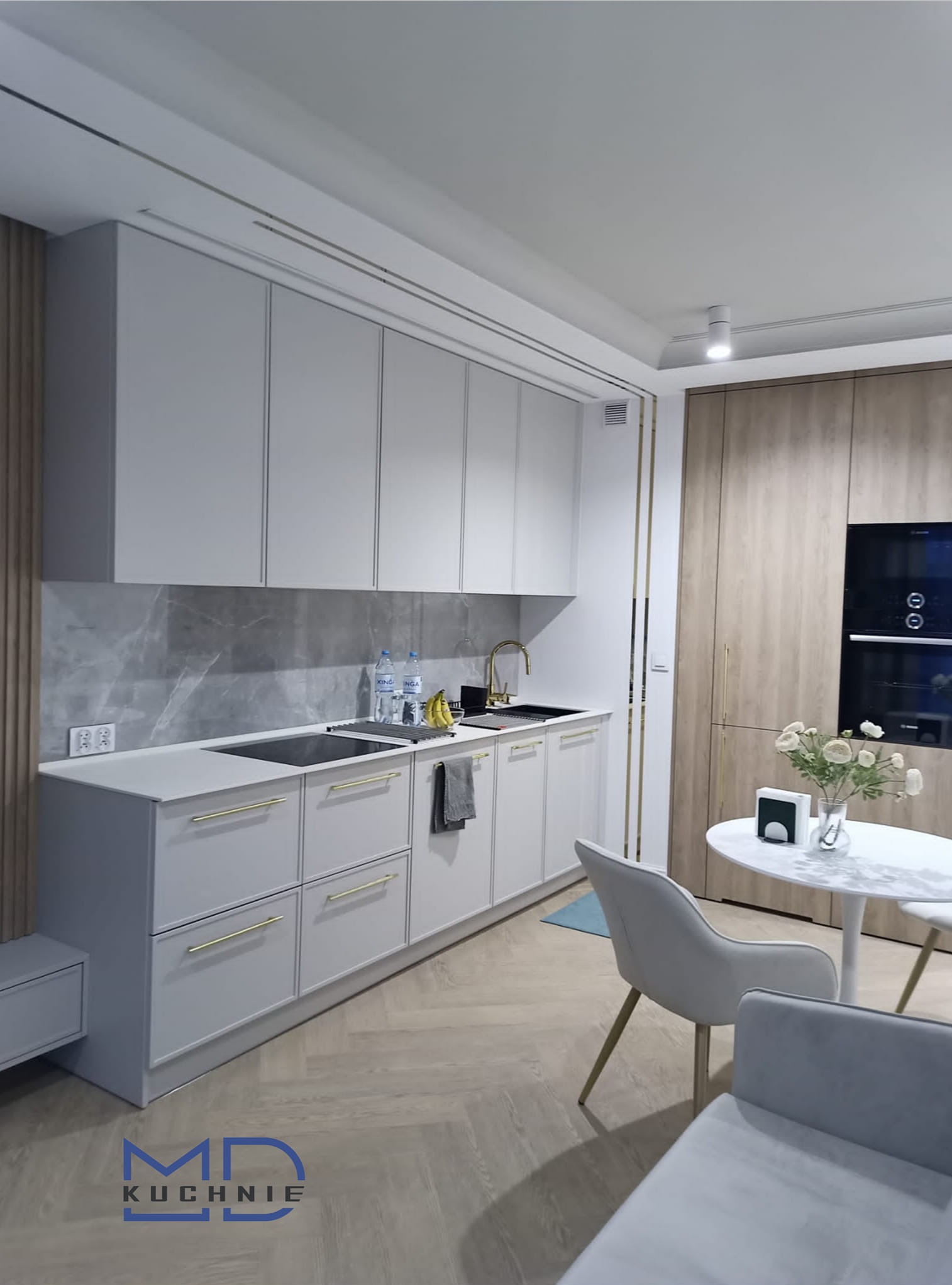 Jasna kuchnia z szarymi frontami, złotymi uchwytami i marmurowym panelem nad blatem. Widoczne butelki z wodą, owoce, złota armatura i drewniana podłoga w jodełkę.