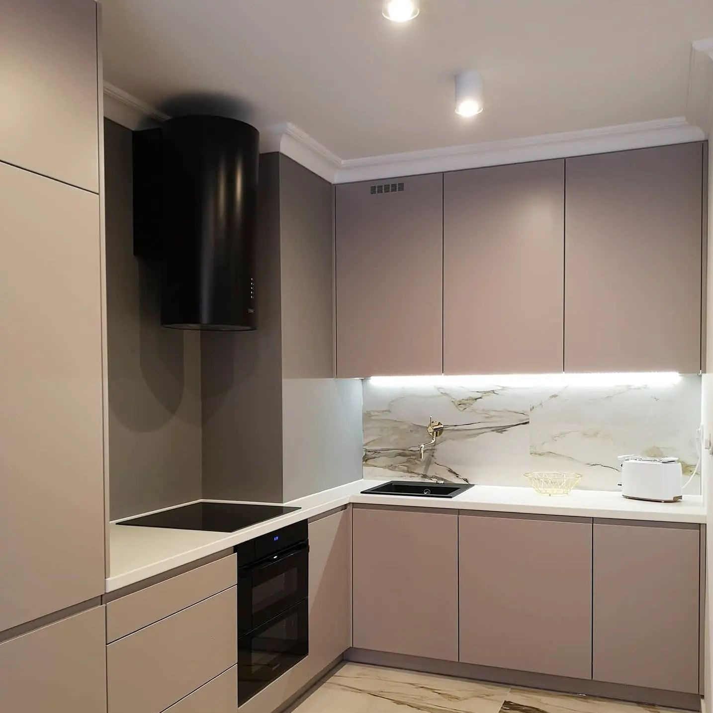 Nowoczesna kuchnia w odcieniach beżu i szarości z cylindrycznym czarnym okapem, marmurowym backsplashiem i minimalistycznym oświetleniem LED pod szafkami.