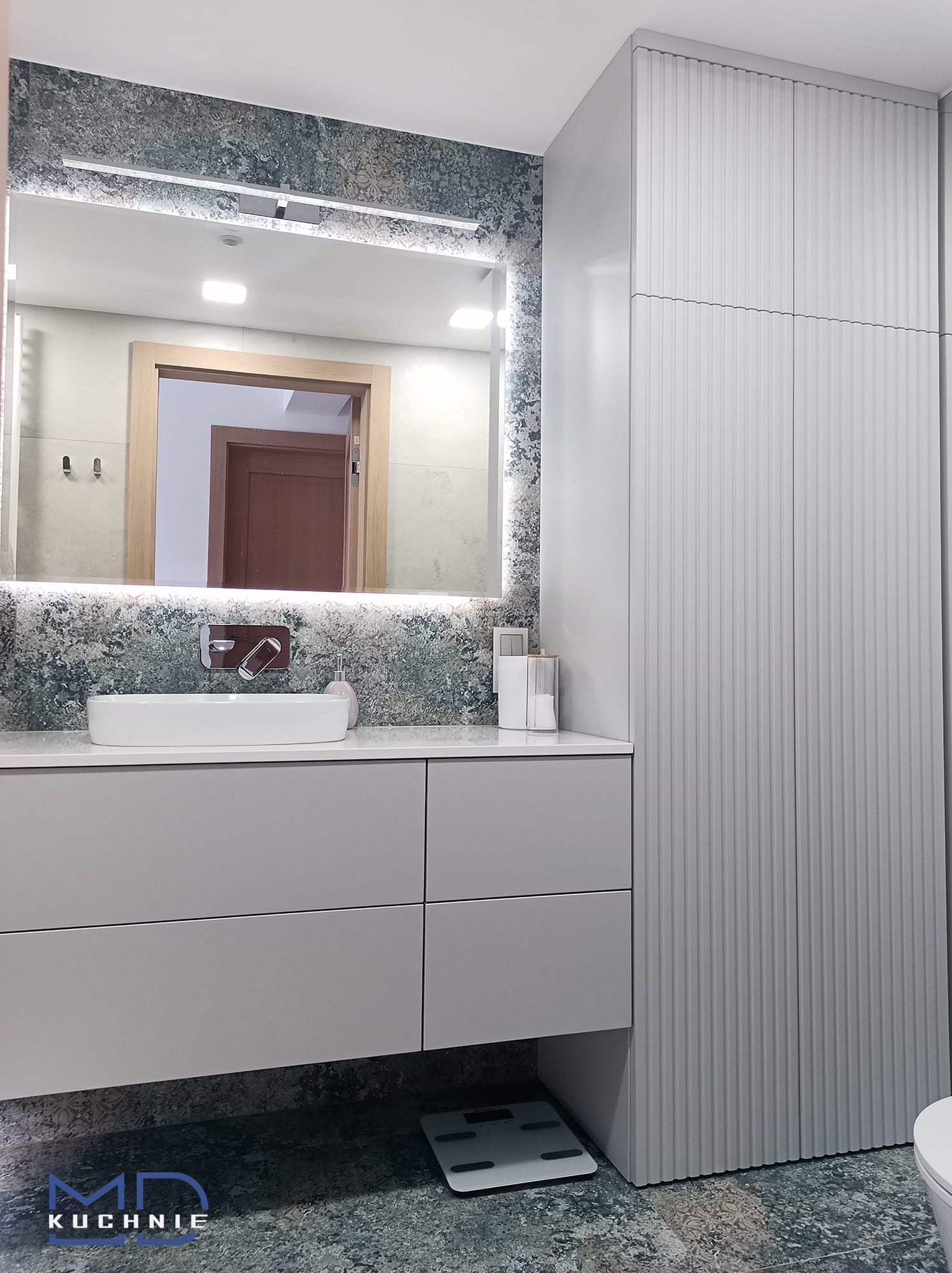 Nowoczesna łazienka z szarą szafką pod umywalkę, umywalką nablatową, podświetlanym lustrem z efektem marmuru na ścianie i wysoką szafą z ryflowanymi drzwiami.