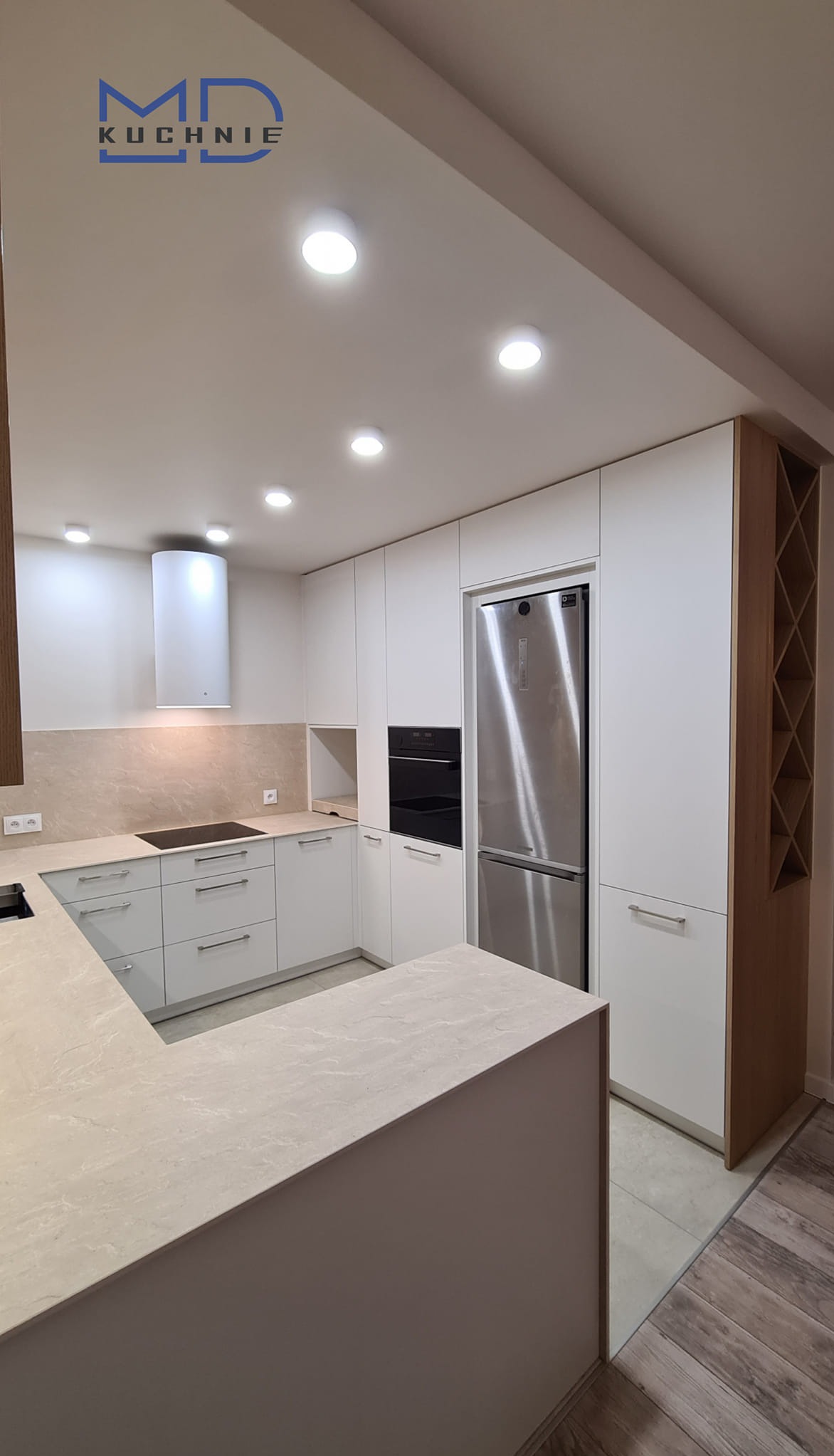 Nowoczesna kuchnia z białymi frontami szafek, beżowym blatem i drewnianym regałem na wino, oświetlona punktowymi lampami sufitowymi.