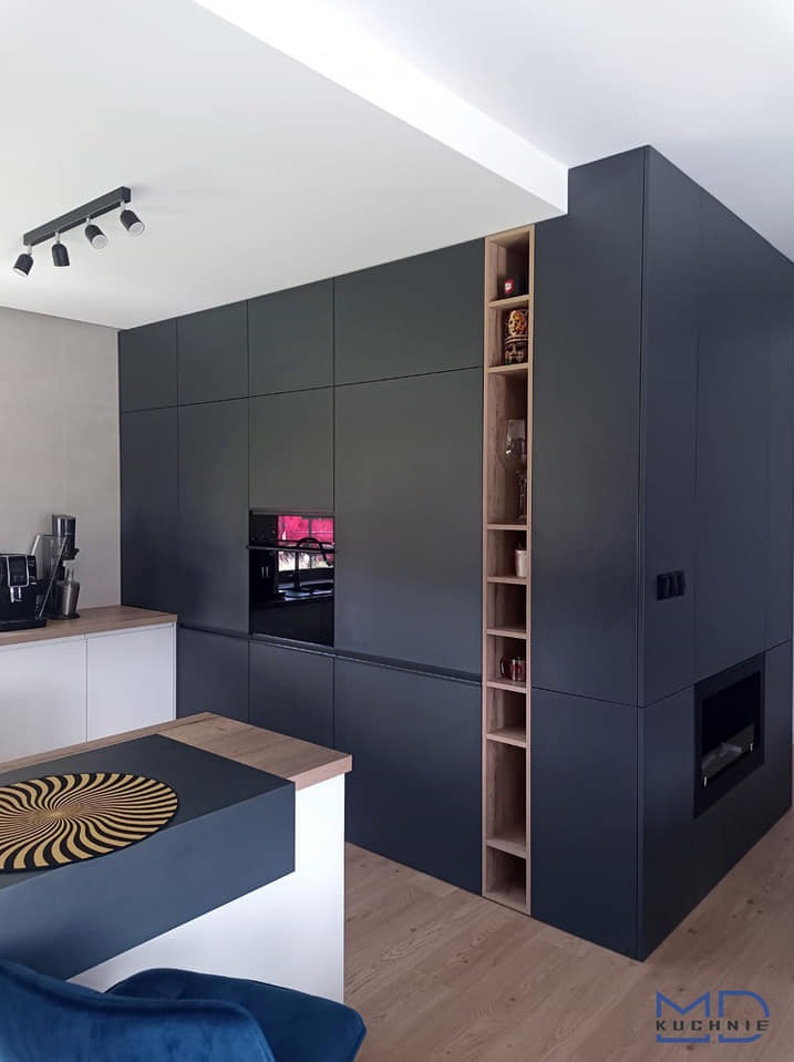 Nowoczesna kuchnia z zabudową w kolorze grafitowym, drewnianymi akcentami w postaci otwartych półek, wyspą z blatem w dwóch kolorach i wbudowanym piekarnikiem.