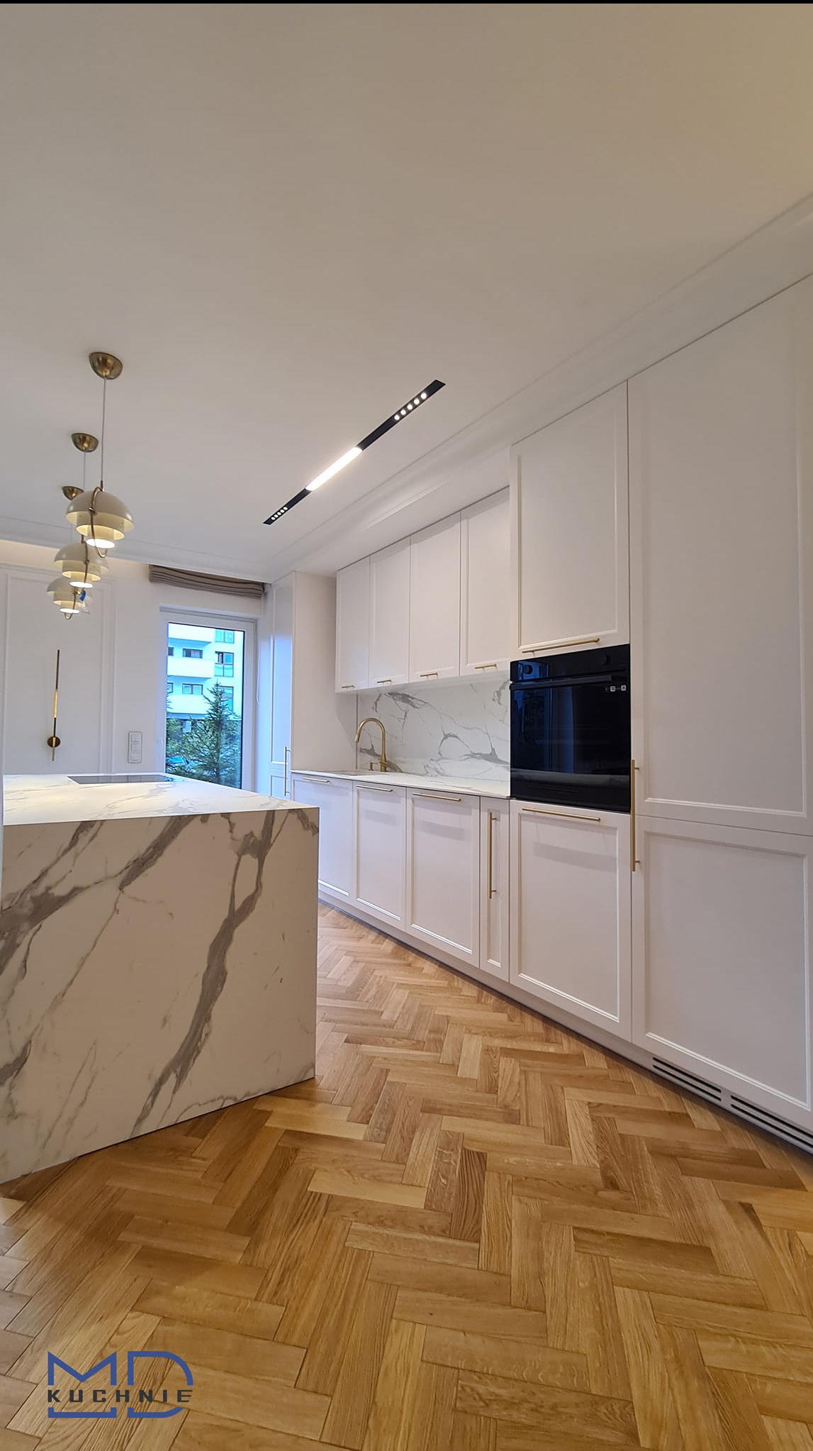 Elegancka kuchnia z białymi frontami szafek, złotymi uchwytami i blatem z marmuru carrara, parkiet w jodełkę, nowoczesne oświetlenie sufitowe.