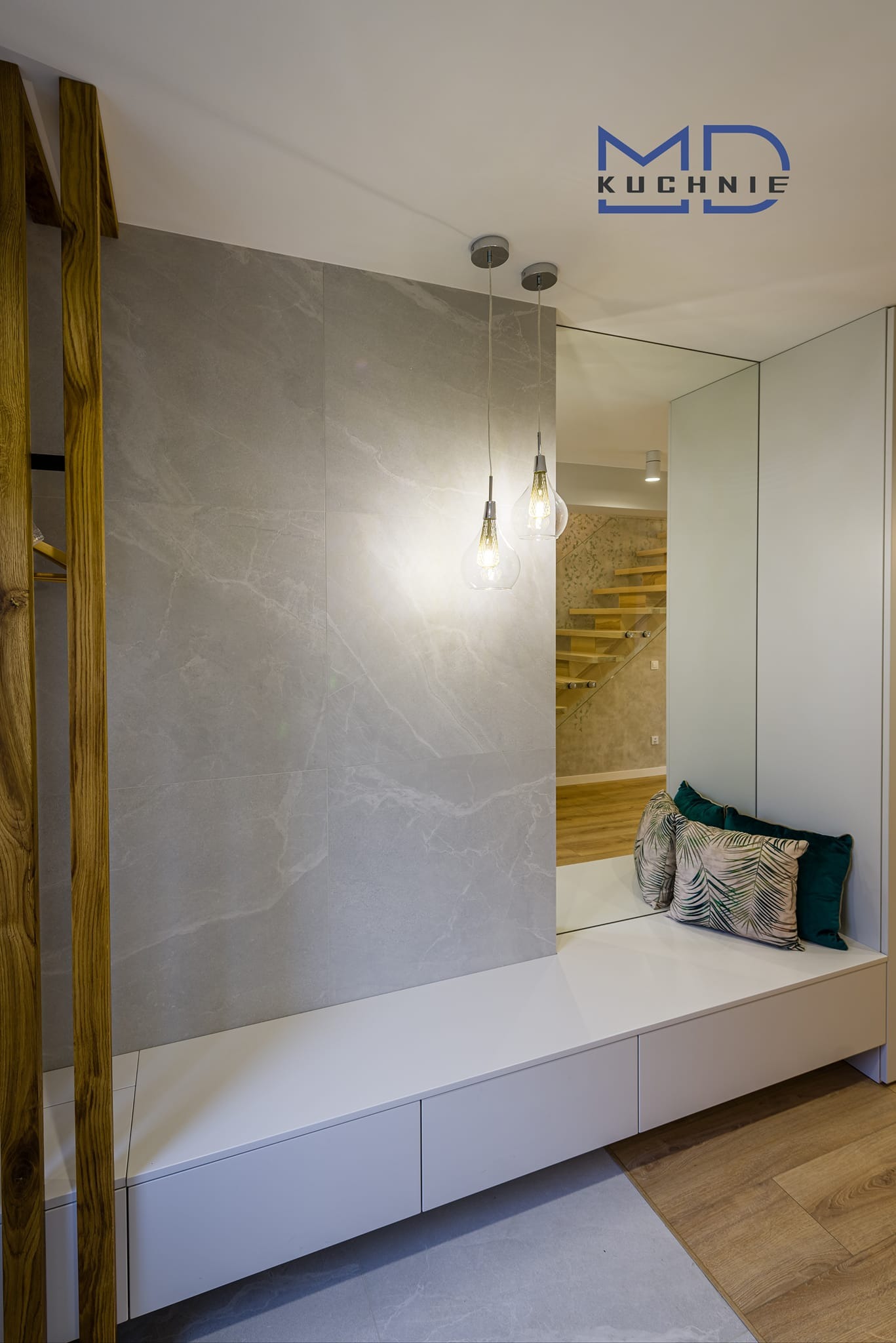 Nowoczesna aranżacja przedpokoju z siedziskiem i szafkami w zabudowie, szara ściana z płytek imitujących marmur, drewniane elementy dekoracyjne, lustro i widok na drewniane schody w tle...