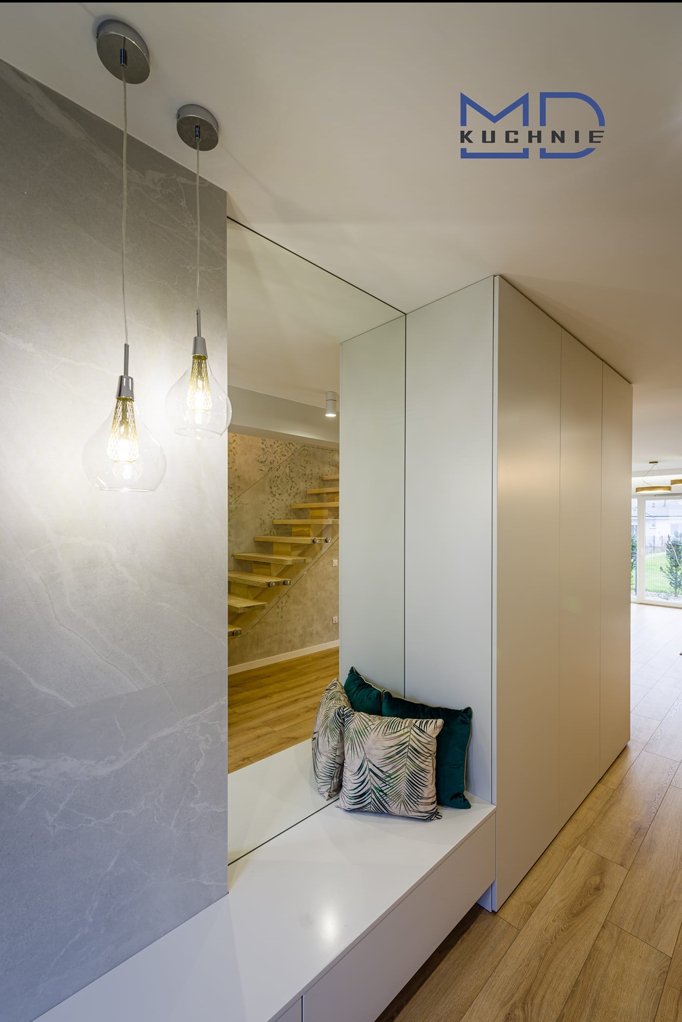 Nowoczesny przedpokój z lustrzanym panelem, wbudowaną szafą z jasnymi frontami i siedziskiem z poduszkami w tropikalne wzory, oświetlony wiszącymi lampami z żarówkami dekoracyjnymi.