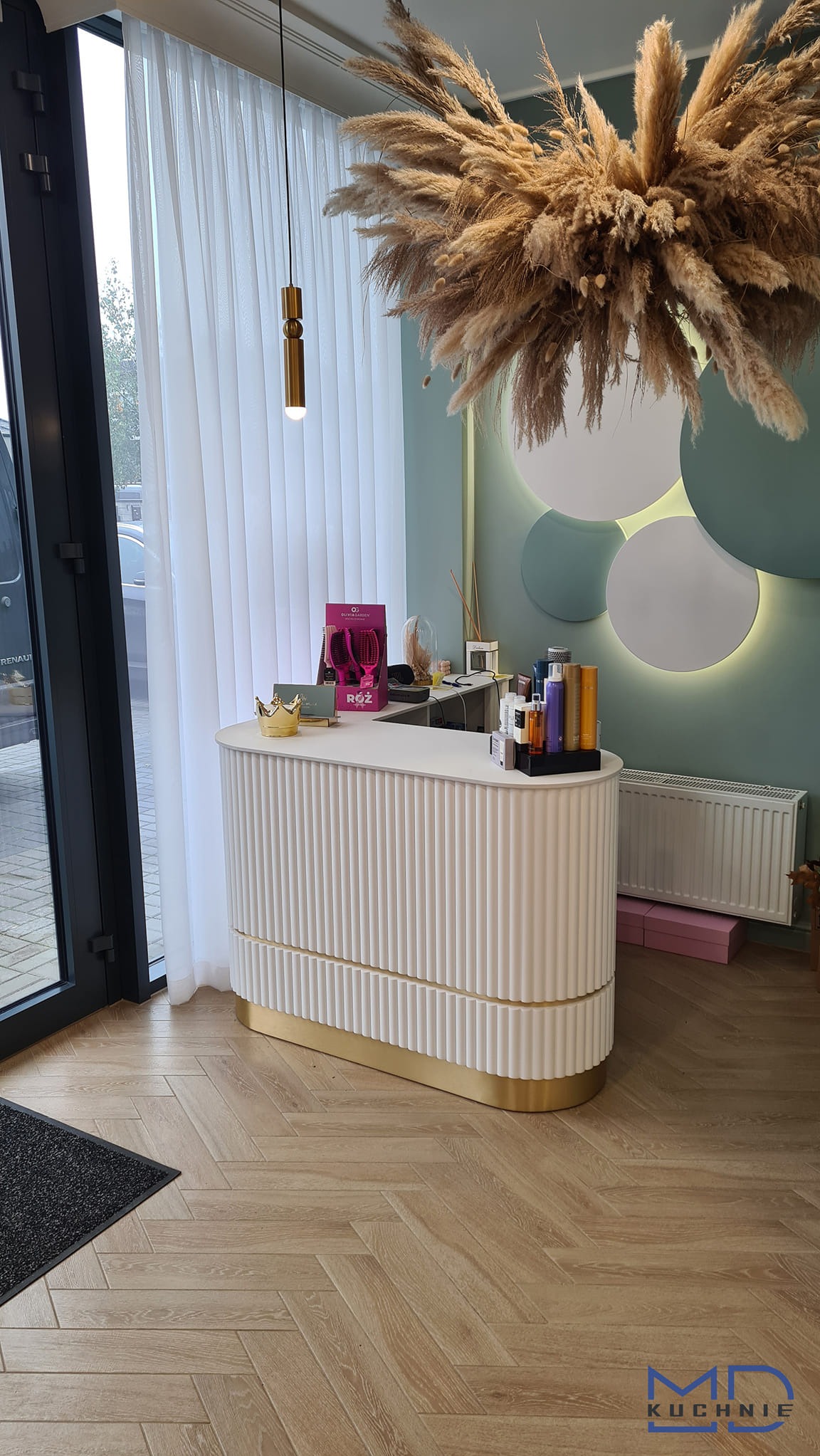 Nowoczesna lada recepcyjna z białymi, pionowymi żłobieniami i złotymi akcentami, dekorowana puszystą kompozycją z traw pampasowych, oświetlona wiszącą lampą i podświetlanymi okręgami na ścianie...