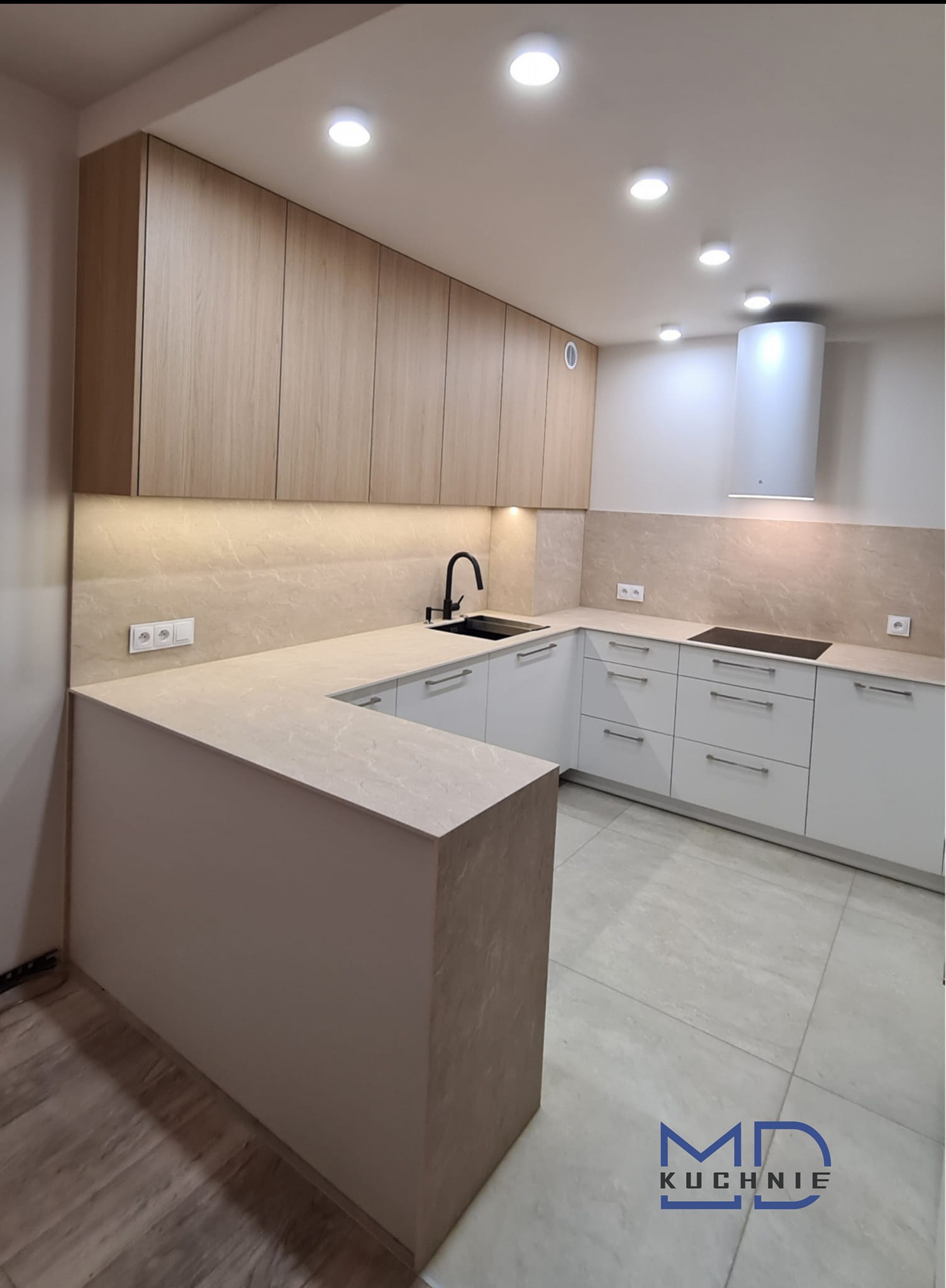 Nowoczesna kuchnia w kształcie litery U z białymi szafkami dolnymi i górnymi szafkami w kolorze jasnego drewna, czarny kran, beżowy blat i backsplash, biały okap kuchenny, widoczne logo firmy...