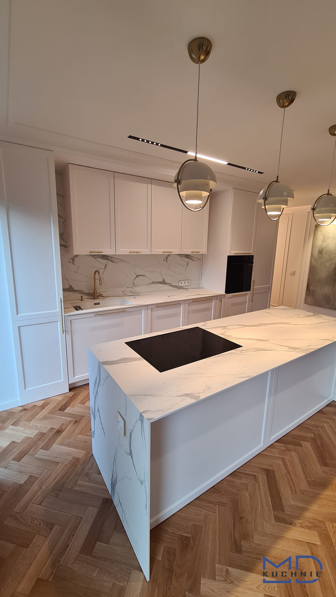 Elegancka kuchnia z białą zabudową, marmurowym blatem wyspy kuchennej z płytą indukcyjną i parkietem ułożonym w jodełkę, oświetlona wiszącymi lampami z elementami w kolorze złota.