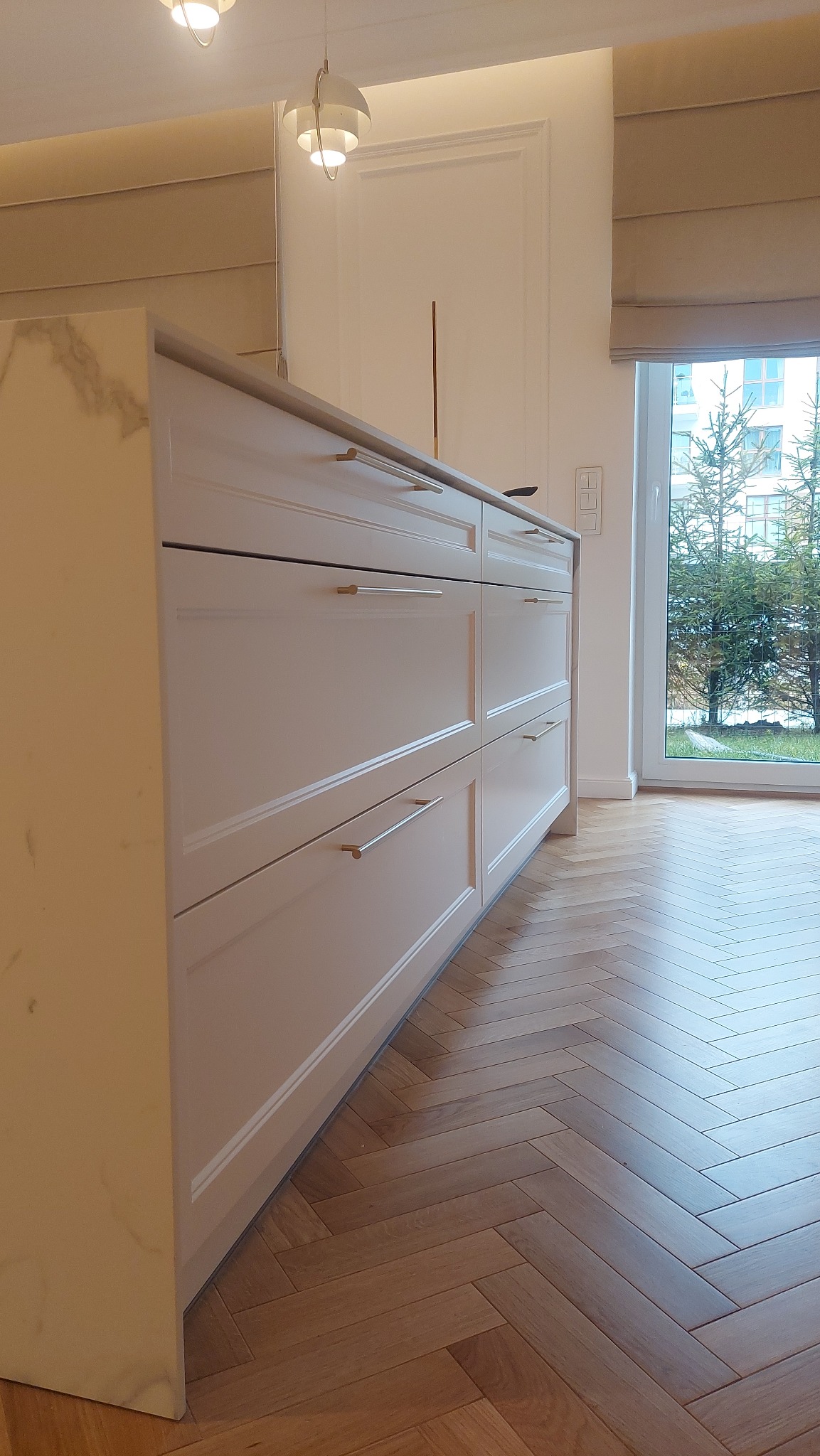 Fragment zabudowy kuchennej z białymi frontami szuflad i marmurowym blatem bocznym, widoczny parkiet w jodełkę i okno z widokiem na zewnątrz.