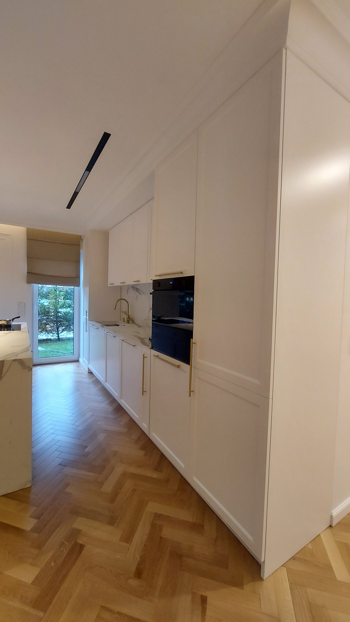 Jasna kuchnia z białymi szafkami, złotymi uchwytami i blatem imitującym marmur, widok z perspektywy podłogi z parkietem w jodełkę francuską.