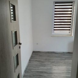 Usługi ogólnobudowlane Krzysztof Jagła - Pusty pokój po remoncie: szare panele podłogowe imitujące drewno, białe ściany, okno z roletami dzień-noc, drzwi wewnętrzne z przeszkleniami.