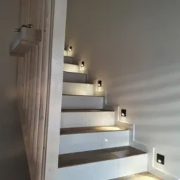 Nowoczesne schody z drewnianymi stopniami i białymi podstopnicami, oświetlone punktowymi lampkami w ścianie, z drewnianą balustradą i doniczką na górze.