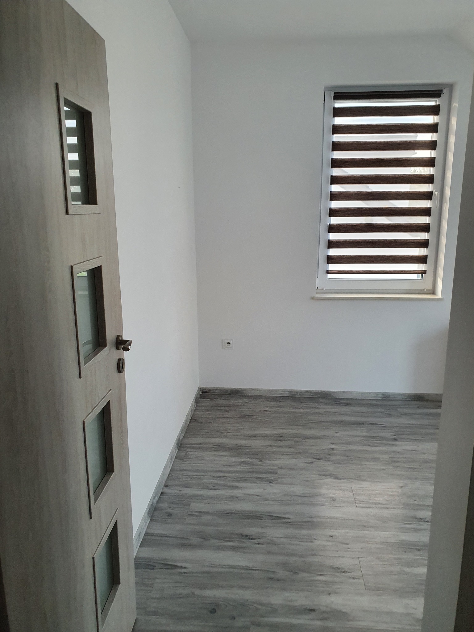 Pusty pokój po remoncie: szare panele podłogowe imitujące drewno, białe ściany, okno z roletami dzień-noc, drzwi wewnętrzne z przeszkleniami.