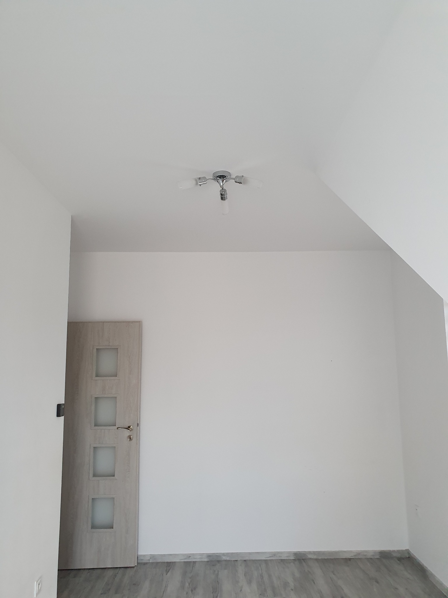 Pusty, świeżo pomalowany pokój z szarymi panelami podłogowymi i drzwiami z czterema przeszkleniami, widok na skośny sufit i nowoczesną lampę sufitową.