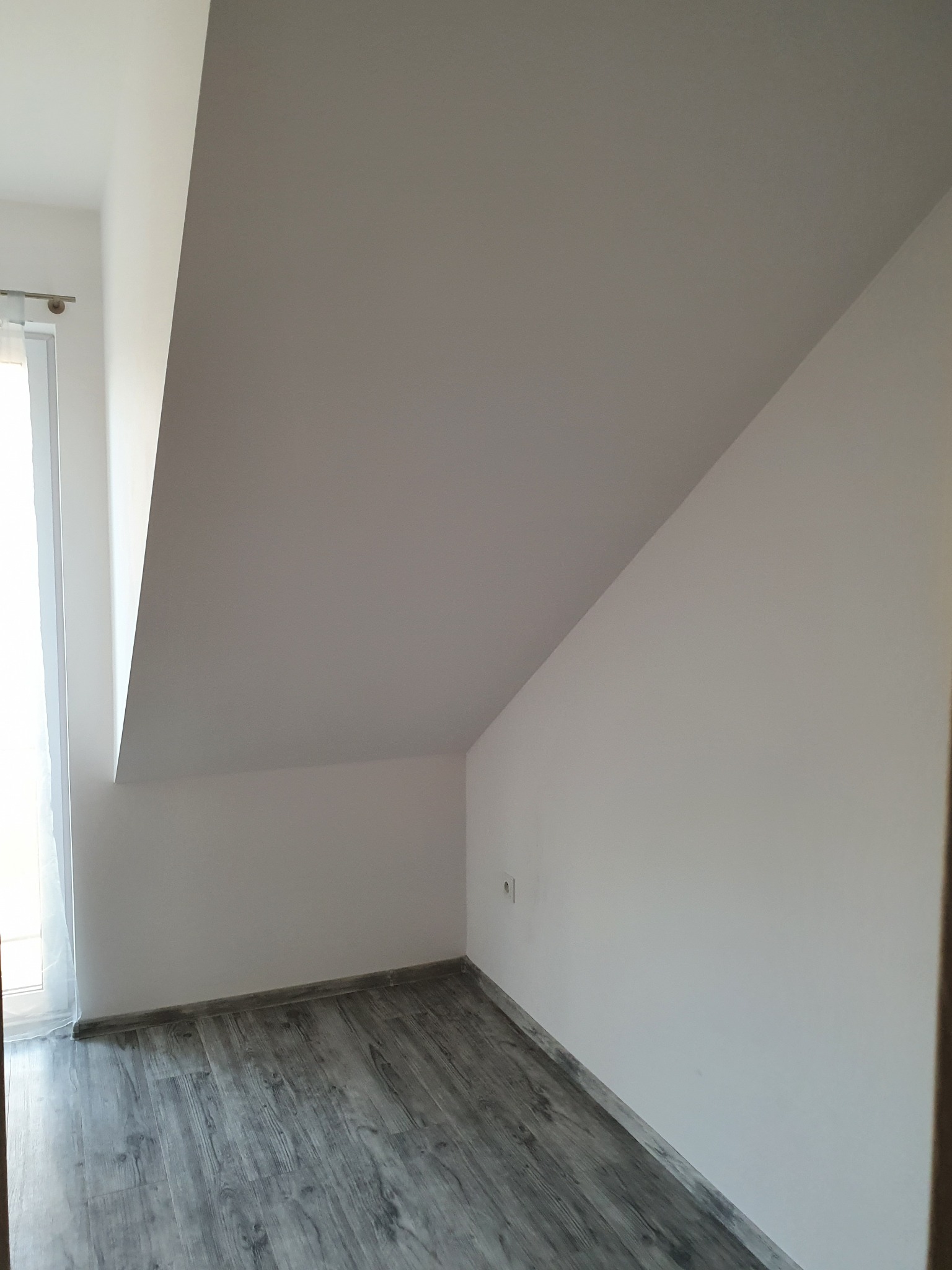 Poddasze po remoncie: szare panele podłogowe imitujące drewno, białe ściany i skośny sufit, widoczne gniazdko elektryczne.