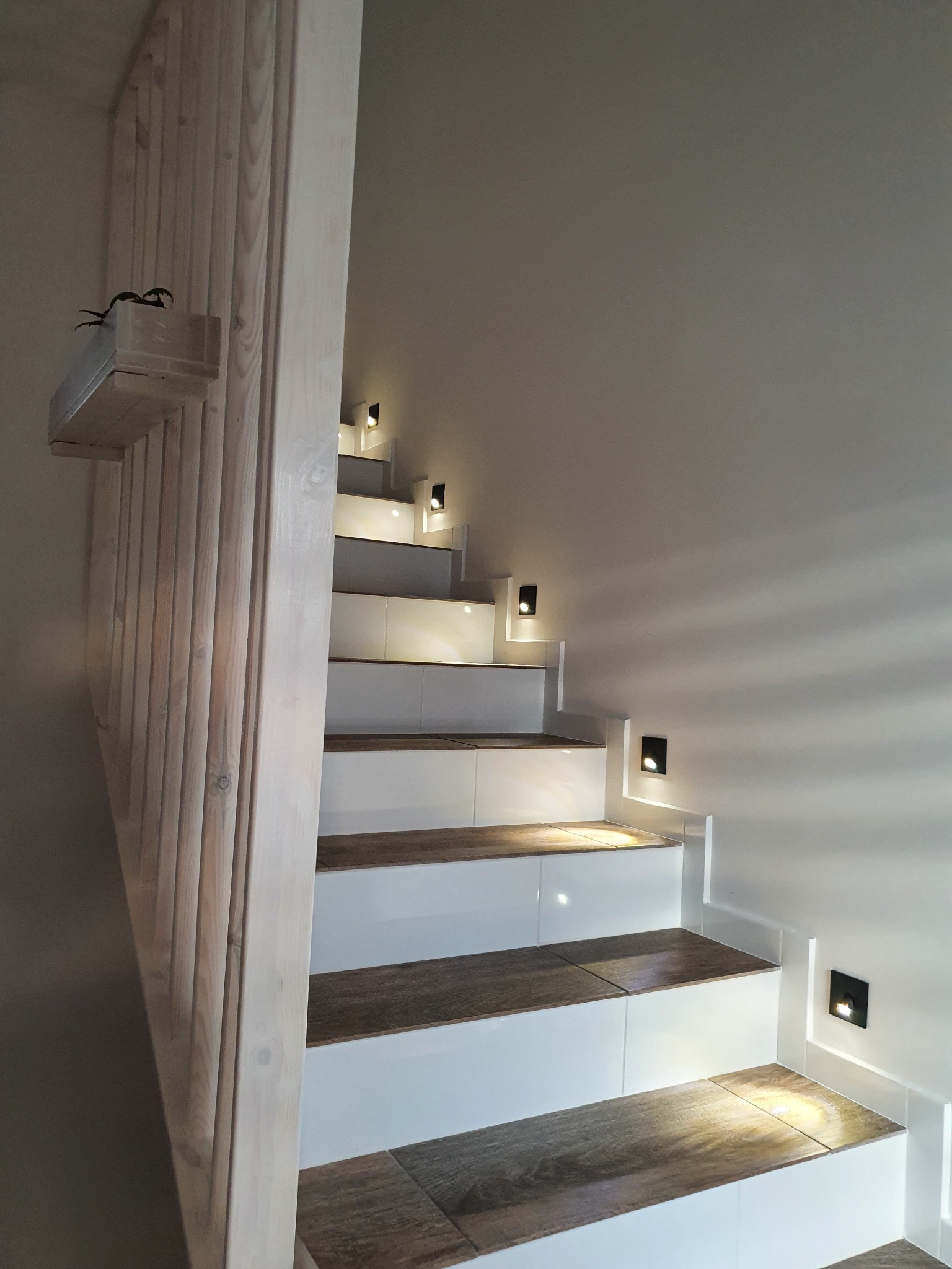 Nowoczesne schody z drewnianymi stopniami i białymi podstopnicami, oświetlone punktowymi lampkami w ścianie, z drewnianą balustradą i doniczką na górze.