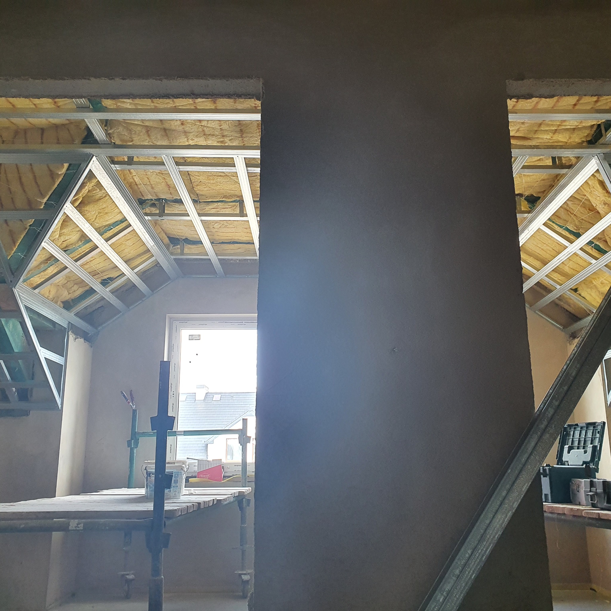 Wnętrze poddasza w trakcie remontu: widoczna konstrukcja metalowa sufitu podwieszanego z izolacją z wełny mineralnej, fragment świeżo nałożonej szarej gładzi na ścianie, rusztowanie robocze...