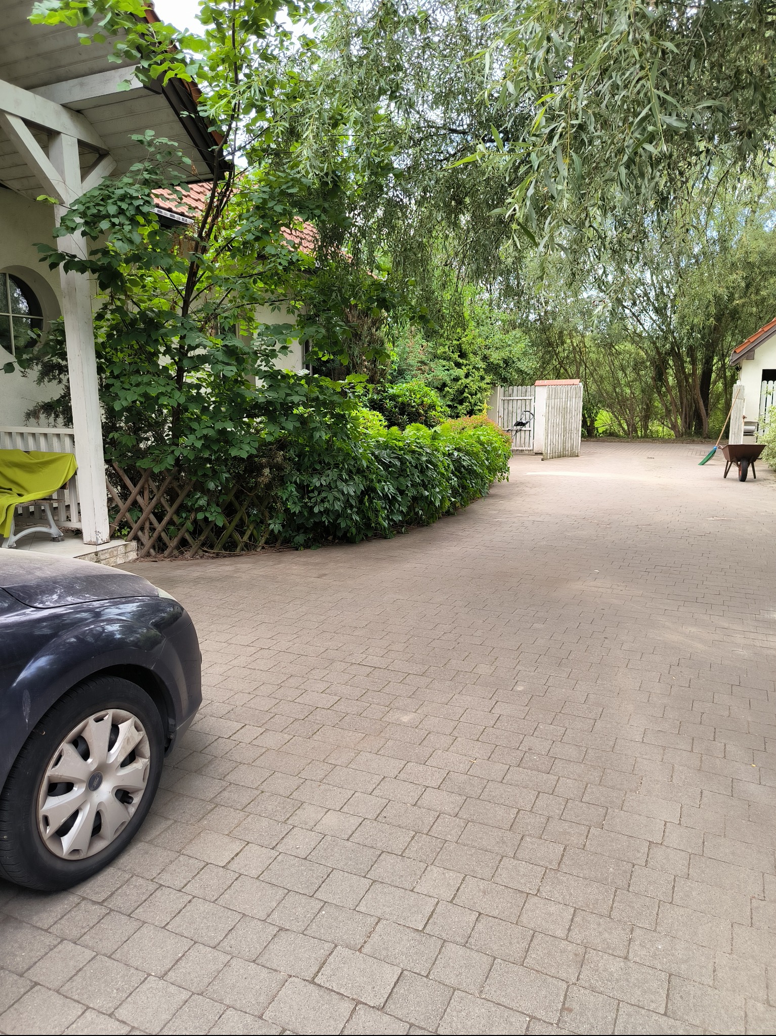 Podjazd wyłożony kostką brukową prowadzący do zadbanego ogrodu z bujną zielenią, widoczny fragment samochodu po lewej stronie, w tle narzędzia ogrodnicze.